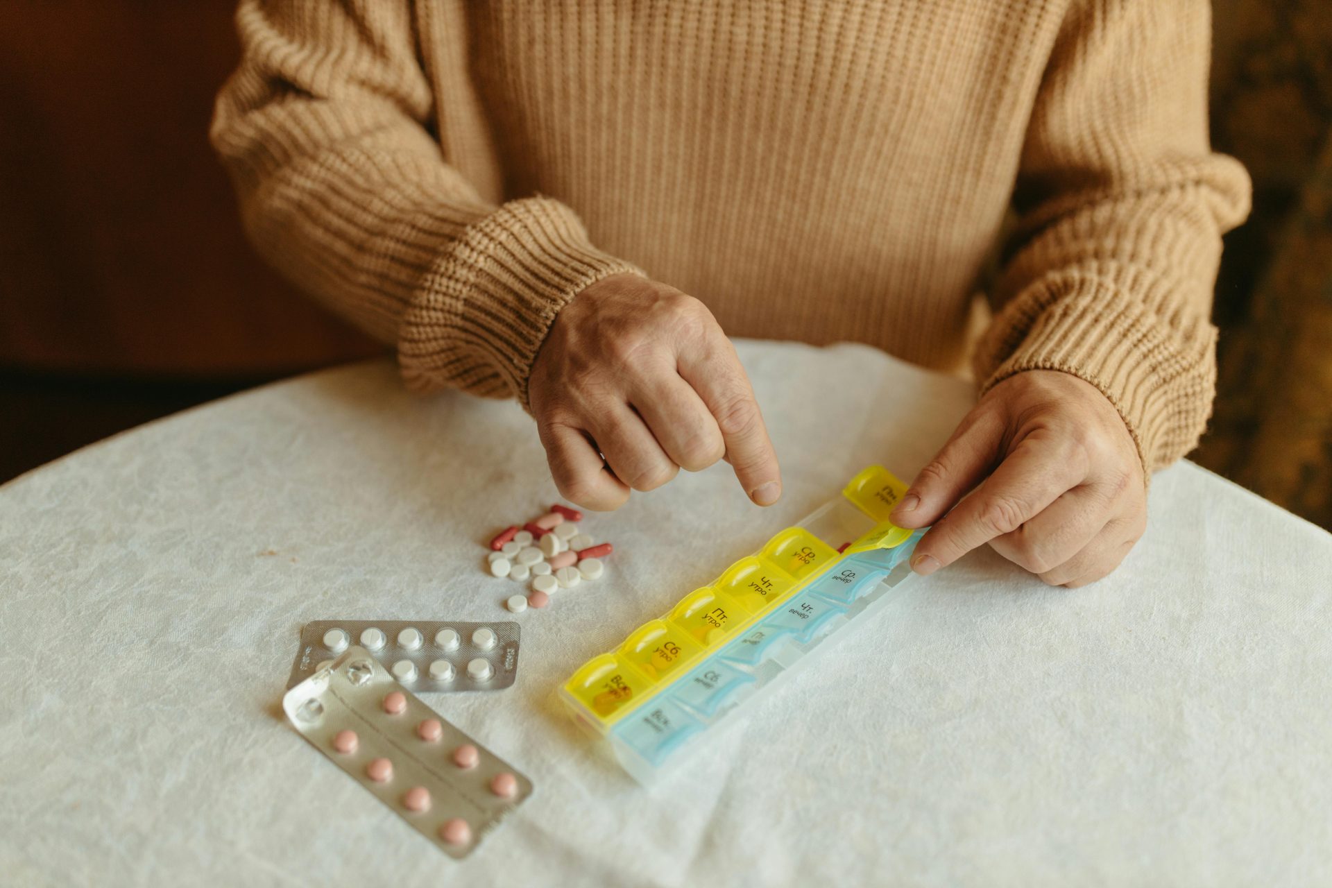 Senior adult organizing daily medications using a weekly pill organizer on a table indoors.